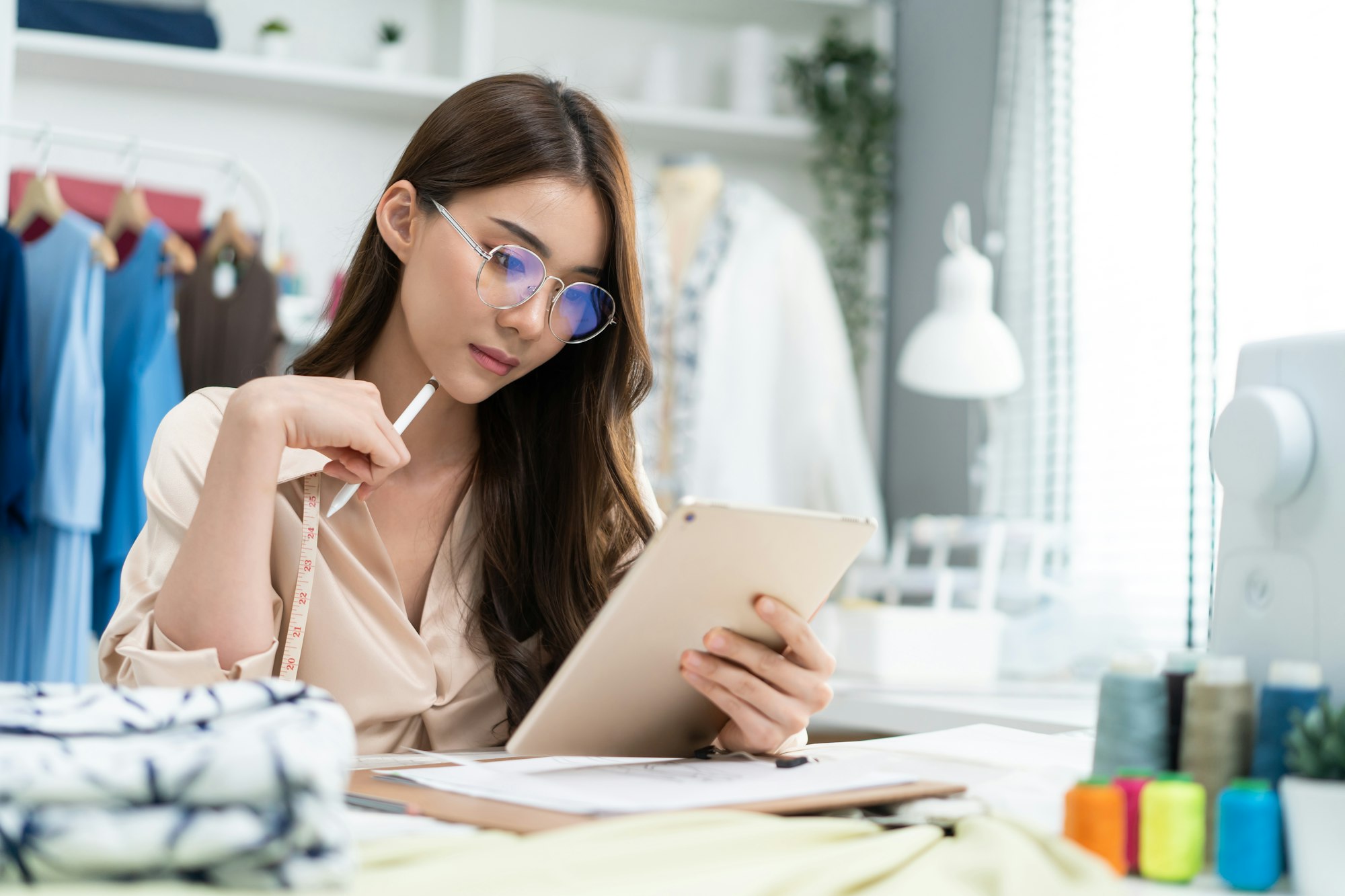 Asian tailor woman use tablet to find reference to think and design new clothes in workshop room